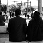 Two graduates in black gowns sitting on a low wall, viewed from behind, watching people at an outdoor event.