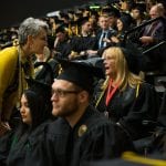 Woman in yellow blazer speaking with a seated female graduate among other graduates during a graduation ceremony.