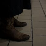 A person wearing brown cowboy boots stands on a tiled floor, with the rest of their outfit mostly obscured in shadow.