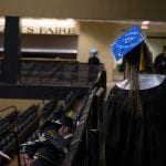 A graduate in cap and gown takes a selfie at a commencement ceremony, with an empty seating area and other graduates in the background.