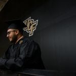 A male graduate in a black cap and gown stands at a podium, looking away from the camera, with a ucf logo on the wall behind him.