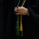 Close-up of a person in a graduation gown holding colorful cords, with a focus on their hands and the texture of the gown.