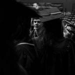 Graduation ceremony in a large hall, depicting a male graduate in cap and gown looking attentive, surrounded by peers in similar attire. black and white photo.