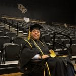 A smiling graduate in a cap and gown sits in an empty auditorium at ucf, holding a diploma.