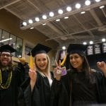 Three graduates in caps and gowns joyfully gesture peace signs at a graduation ceremony.