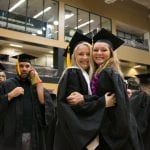 Two smiling female graduates hugging at a graduation ceremony, surrounded by other graduates in caps and gowns.