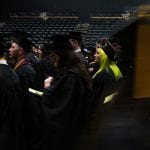 Graduates in caps and gowns walking in procession during a dimly lit college graduation ceremony.