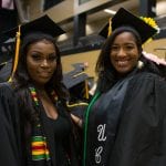 Two female graduates in black robes and caps, smiling at a graduation ceremony, adorned with cultural sashes.
