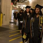 Two graduates in black robes and decorated caps walking happily in a hallway during a graduation ceremony.