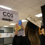Graduate in a cap and gown takes a selfie with a sign reading "commencement cos college of sciences bachelor's candidates" at a university ceremony.