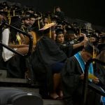 Graduates wearing black gowns and decorated caps seated in stadium seating at a commencement ceremony.