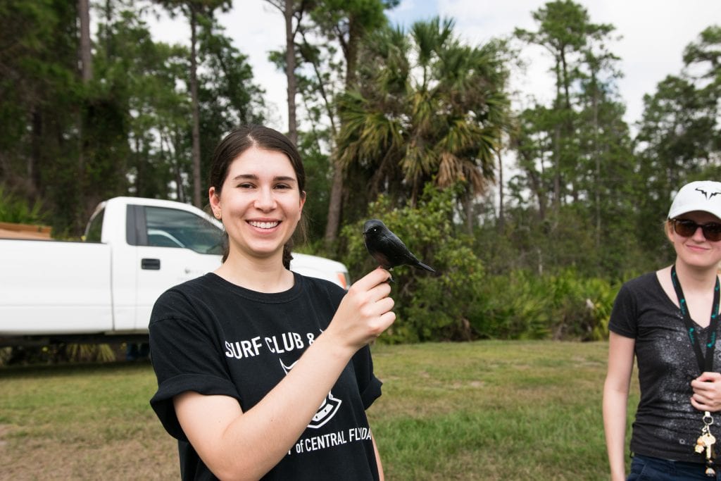 A smiling woman in a surf club t-shirt holds a small blackbird outdoors, with another person partially visible in the background.