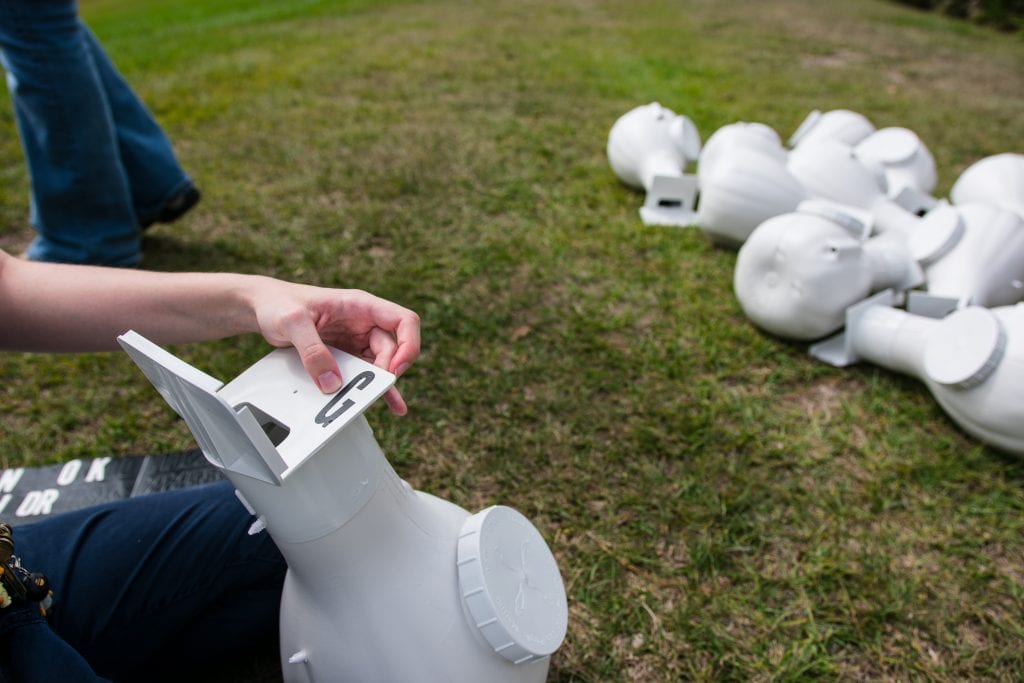 A person inserting a battery into the head of a white, humanoid robot lying on grass, with other disassembled robot parts nearby.