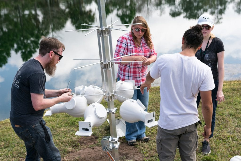 Group of four people setting up scientific equipment by a lake, with one person taking notes.