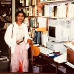 A woman stands in an office, holding books, surrounded by bookshelves, file cabinets, and old computers.