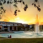 Two people sitting by a fountain in a park at sunset, with modern buildings in the background and trees framing the scene.