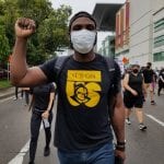 Man wearing a mask and a black t-shirt raises his fist during a protest, with people and buildings in the background.