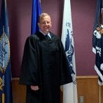 A person in a judge's robe smiling, standing in front of various military flags in an indoor setting.