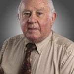 Elderly man in a beige shirt and patterned tie, looking directly at the camera with a neutral expression, set against a gray background.