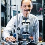 A scientist with a mustache, wearing a white lab coat, adjusts a microscope in a high-tech laboratory filled with electronic equipment.