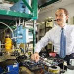 A scientist in a tie works with complex electronic equipment in a lab setting, adjusting components and observing results on a screen.