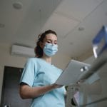 A female nurse wearing a surgical mask and scrubs reviews a patient's records on a digital tablet in a brightly lit hospital room.