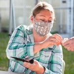 A woman in a plaid shirt and mask receives a card from a hand while seated at an outdoor table, holding a phone.