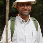 Man in a jungle wearing a white hat and glasses, holding a wooden stick, with a backpack, smiling at the camera.