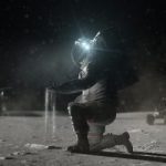 Astronaut kneeling and collecting soil samples on a dusty, moon-like surface at night, illuminated by helmet lights, with a rover in the background.