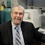 A smiling, bearded man in a suit and tie standing in a laboratory with equipment in the background.