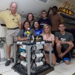 A diverse group of eight people posing with a scientific experiment setup inside a nasa aircraft cabin.