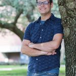 Man in glasses smiling, leaning against a tree with arms crossed, wearing a blue shirt and jeans in a park.