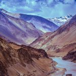 A panoramic view of a winding river flowing through a rugged mountain valley with varied hues of brown and purple, capped with snowy peaks.