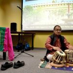 A man in traditional attire playing tablas beside a projector screen displaying a building, with shoes off and a colorful cloth on the floor.