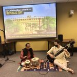 Two men performing traditional indian music, one playing the sitar and the other the tabla, in front of a screen displaying "a celebration of india's republic day.