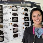 A woman with dark hair, smiling, wearing a pink respirator, stands next to a wall chart displaying various mineral samples.
