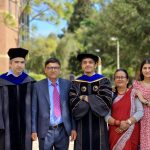 Two graduates in robes with three family members, standing outside on a sunny day.