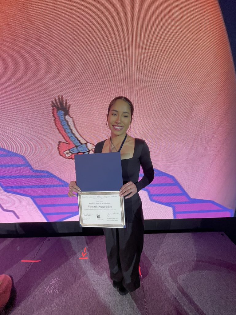 Stefannia Tasayco on stage holding a folder with her award