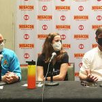 Three people sitting at a table with microphones at a megacon orlando panel, one wearing a star trek costume, all wearing masks.