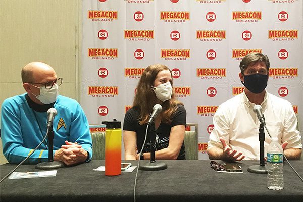 Three people sitting at a table with microphones at a megacon orlando panel, one wearing a star trek costume, all wearing masks.