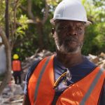 An african american construction worker with a beard, wearing a white helmet and orange safety vest, stands in front of a debris-laden background.