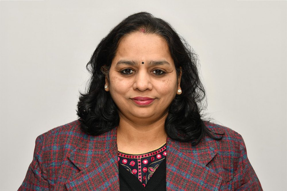 Portrait of Ruchita Gupta in a plaid blazer, smiling against a plain background.