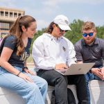 Three people are sitting outdoors, looking at a laptop. One is wearing a white cap and jacket, one has a gray shirt, and one is in a black t-shirt. A building and trees can be seen in the background.