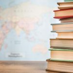 A stack of books is placed on a wooden surface with a blurred world map in the background.