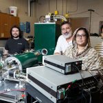 Five people stand in a lab with various scientific equipment, including control panels, monitors, and machinery.