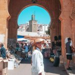 A person wearing a hat is walking through an archway in a bustling outdoor market, with various vendors and shoppers around. A tall structure is visible in the background.