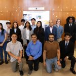 A group of 15 people, of diverse backgrounds, posing for a photo in front of research posters displayed on a wooden wall. They appear to be in a formal setting, with some people standing and others kneeling.