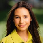 A young woman with long brown hair smiles slightly while looking at the camera. She is wearing a yellow shirt and standing outdoors with a blurred, green background.