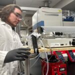 A scientist in protective gear operates a laboratory furnace setup, surrounded by various equipment.