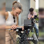 A woman adjusts a camera while a man operates another camera in the background.